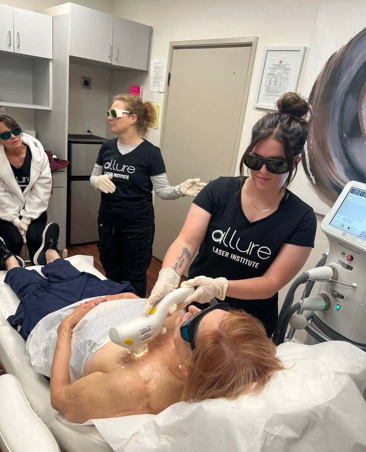 A student in safety goggles applies a laser handpiece to a model's chest while classmates watch from the doorway
