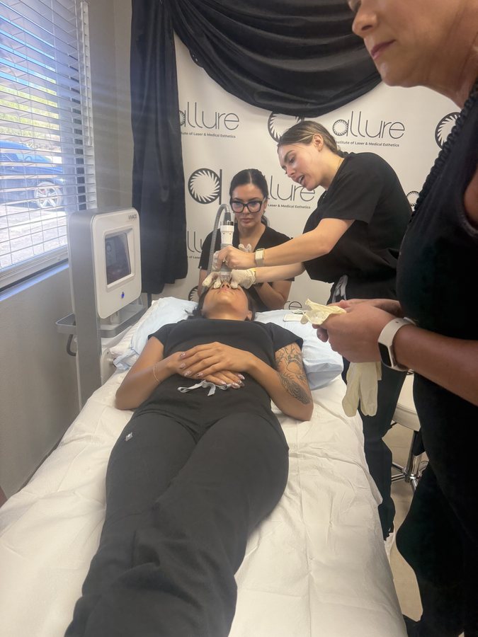 Three people in black shirts treat a model's face with a handpiece as the model lies on a treatment bed