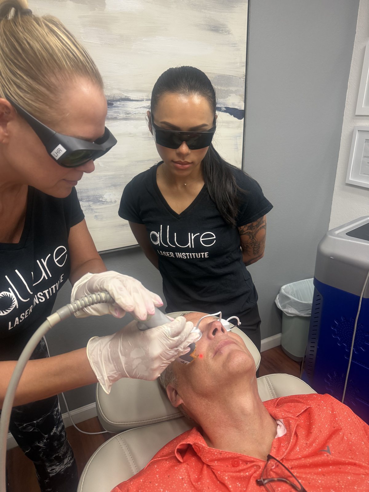 An instructor and a student in laser goggles guide a laser handpiece over a model's face
