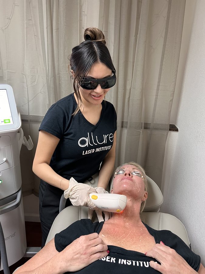 A student in dark safety goggles holds a laser handpiece to a model's neck as the model reclines in a treatment chair