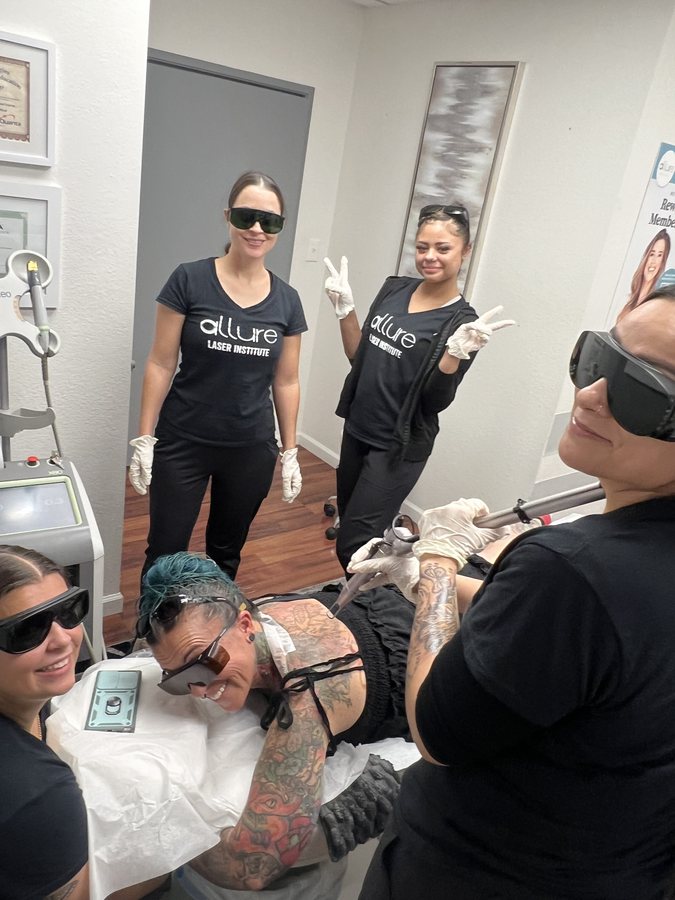 Four people in Allure Laser Institute shirts, goggles and gloves around a treatment bed as a laser handpiece is held over a tattooed model's shoulder