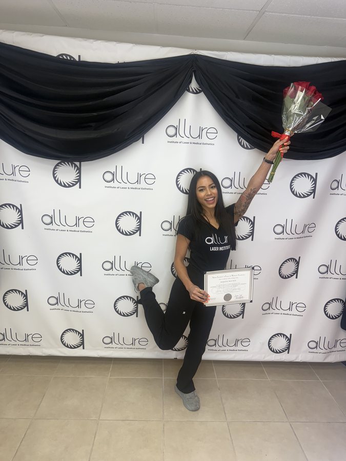 A graduate holding a certificate and a bouquet of roses, one heel kicked up, in front of a white backdrop