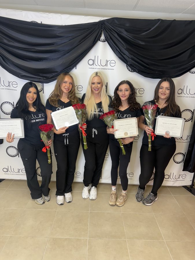 Five graduates in black shirts holding certificates and red roses in front of a black-draped backdrop