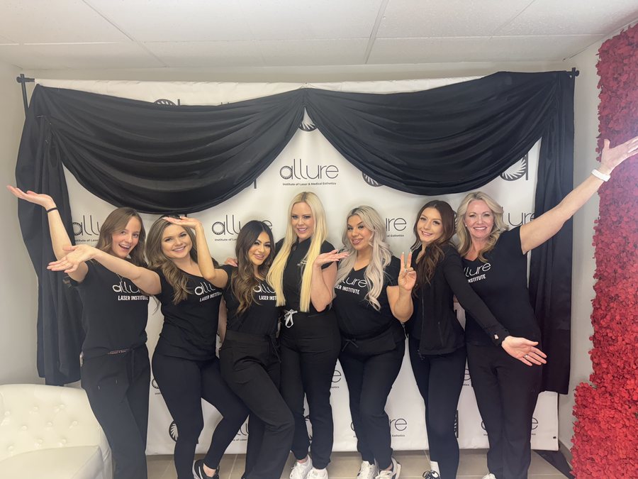 Seven people in black Allure Laser Institute shirts pose with arms out in front of the white Allure step-and-repeat backdrop